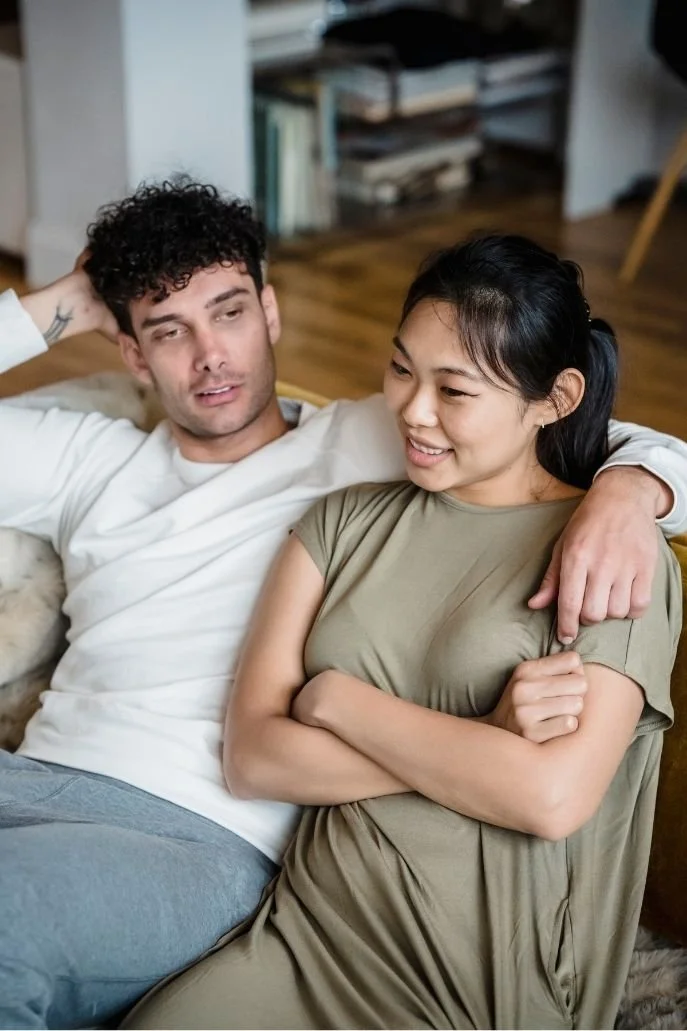 A man and woman sitting on a couch, with the man resting his arm around the woman's shoulders, in a cozy living room.