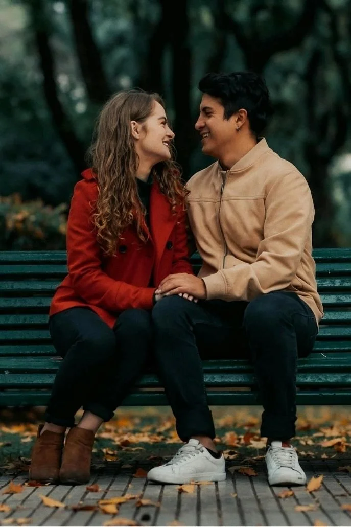 A young couple sitting on a park bench, looking at each other and smiling, surrounded by fall leaves and trees.