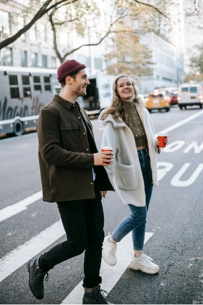 A young man and woman walking on a city street holding red cups, smiling, with cars and buildings in the background during daytime.
