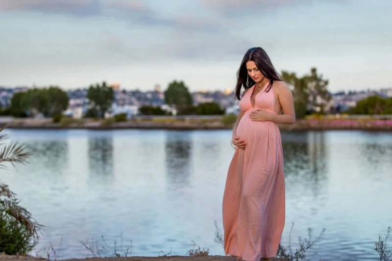 Pregnant woman in a pink dress standing by a lake, looking down at her belly.