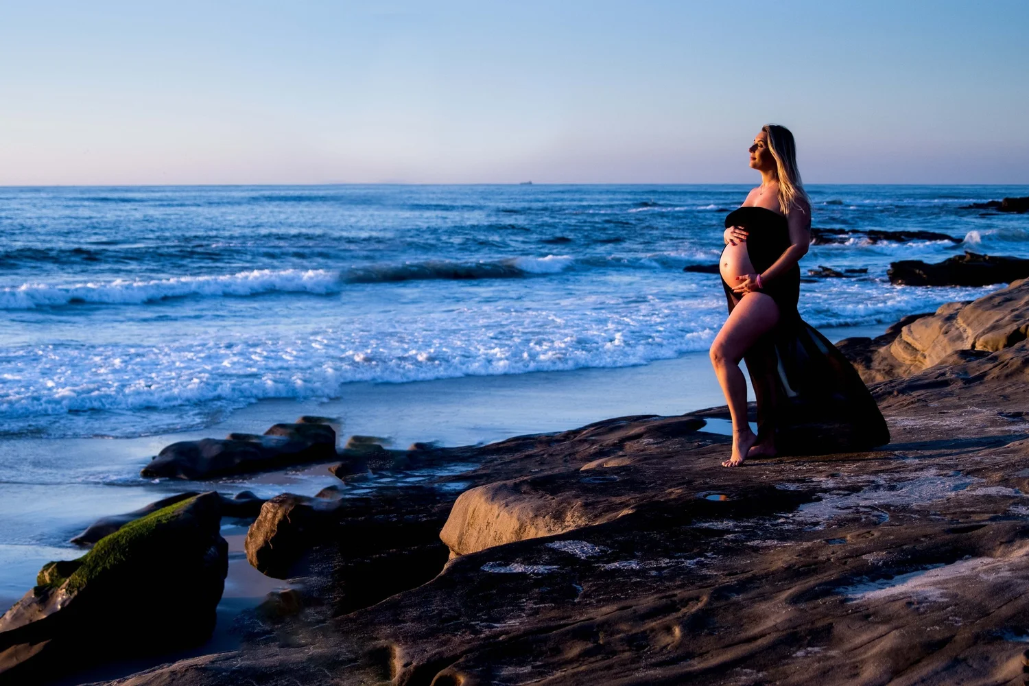 Pregnant woman in a black dress on beach rocks at sunset