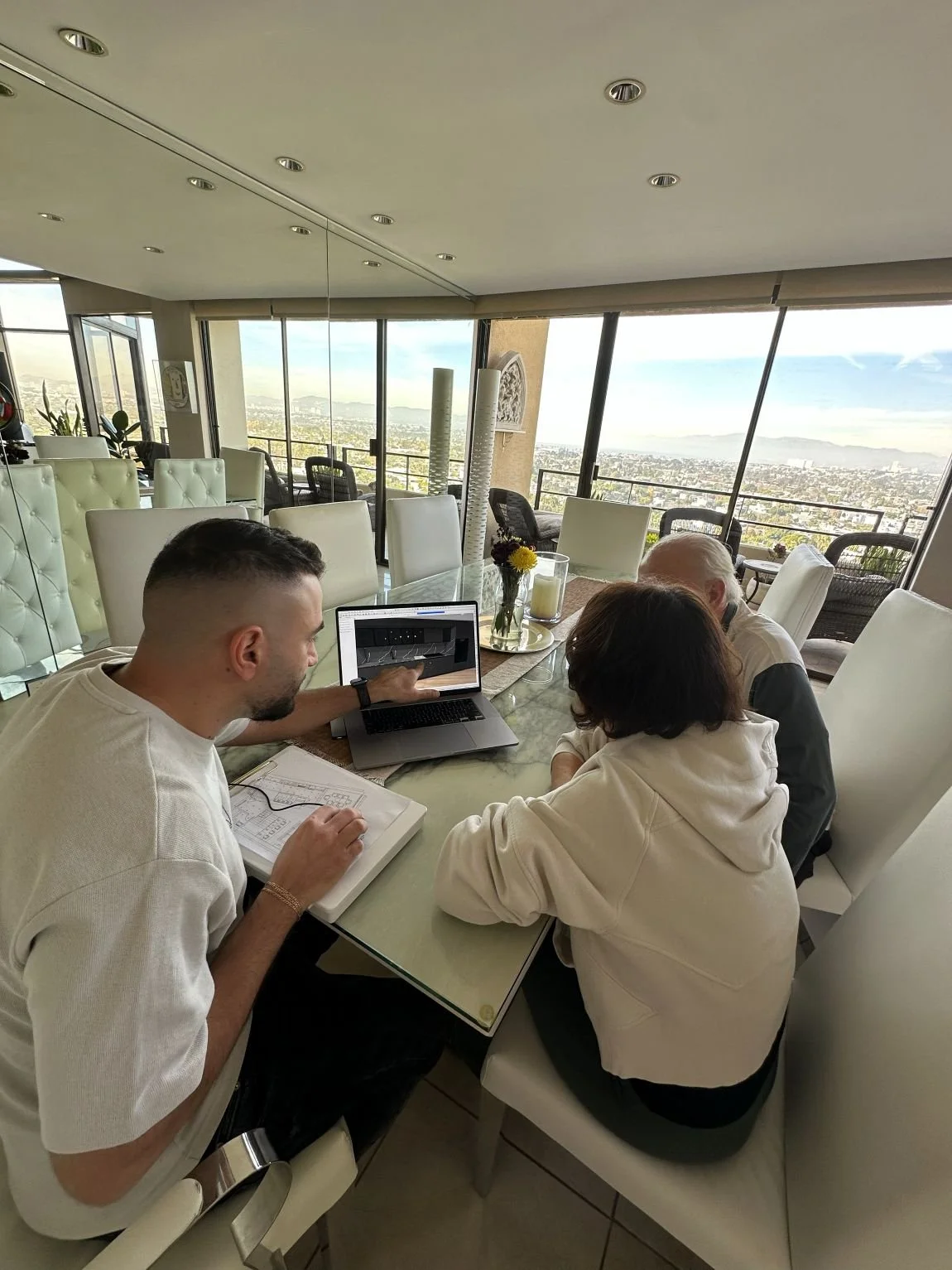 Three people sit at a dining table in a bright room with city views, discussing 3D interior design Los Angeles on a laptop.