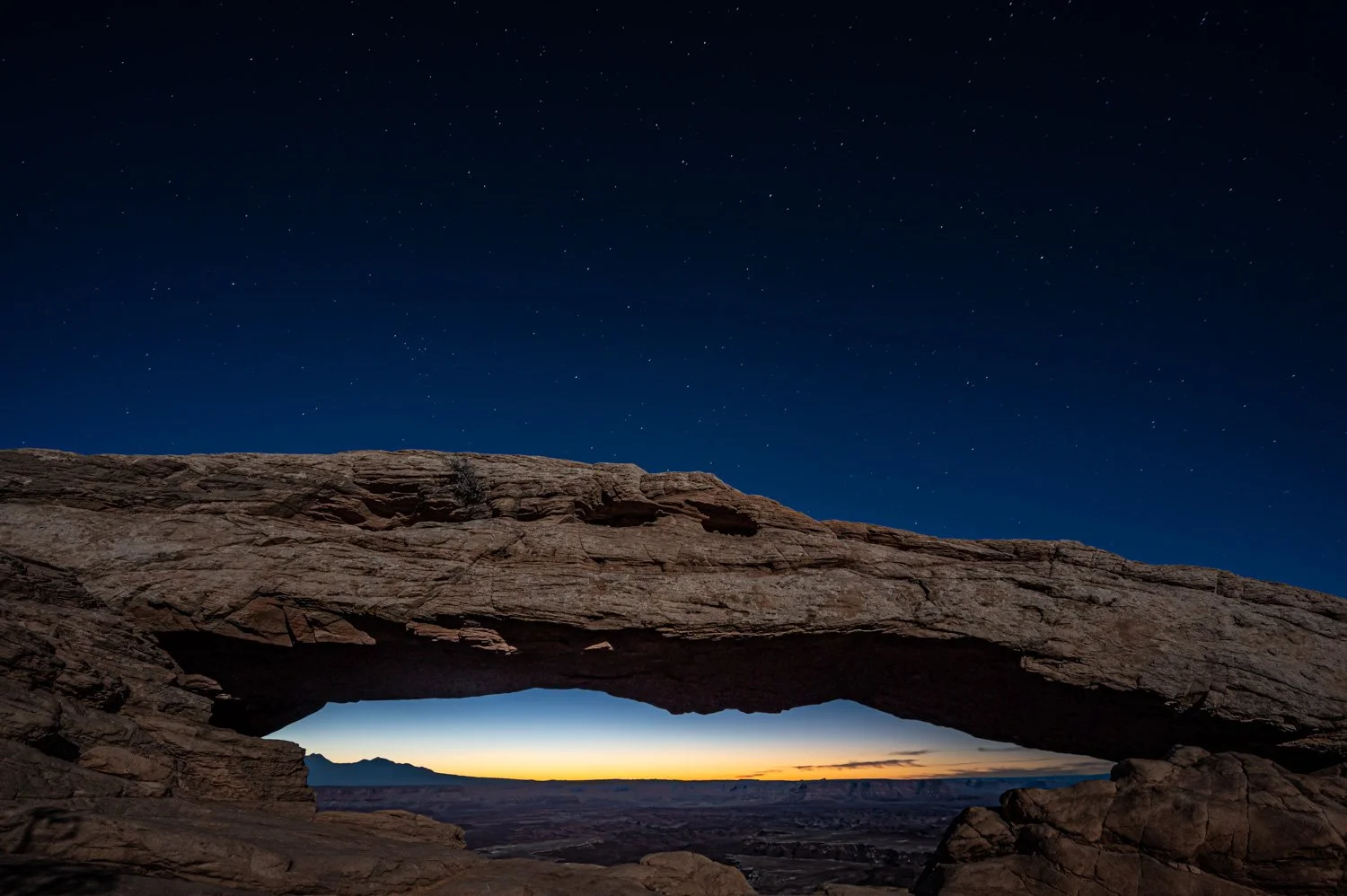 Night view of a natural rock arch with a sunset or sunrise in the distance, starry sky above, and mountain landscape below.
