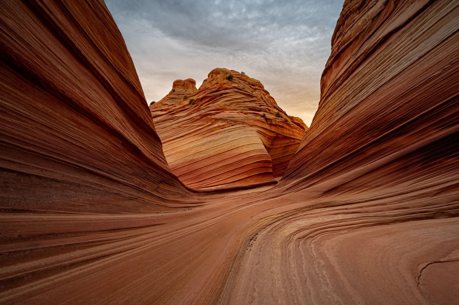 A narrow slot canyon with smooth, layered red and orange rock walls, and a distant hill under a cloudy sky at The Wave in Arizona.