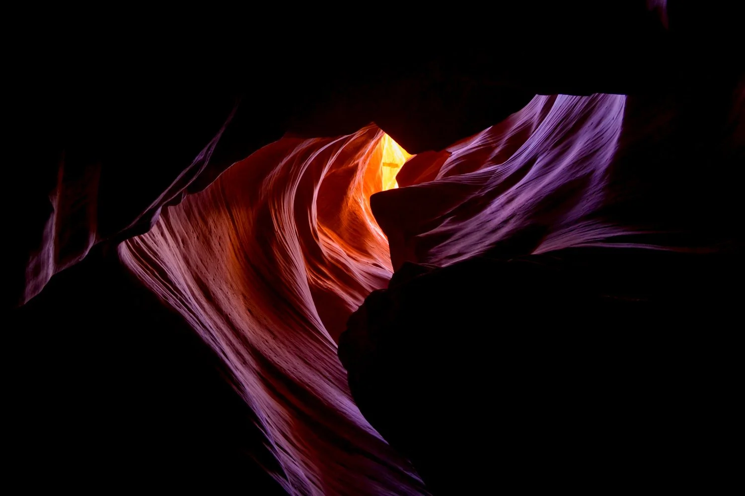 View of a slot canyon with smooth, layered rock formations illuminated by sunlight, creating warm orange and purple hues.