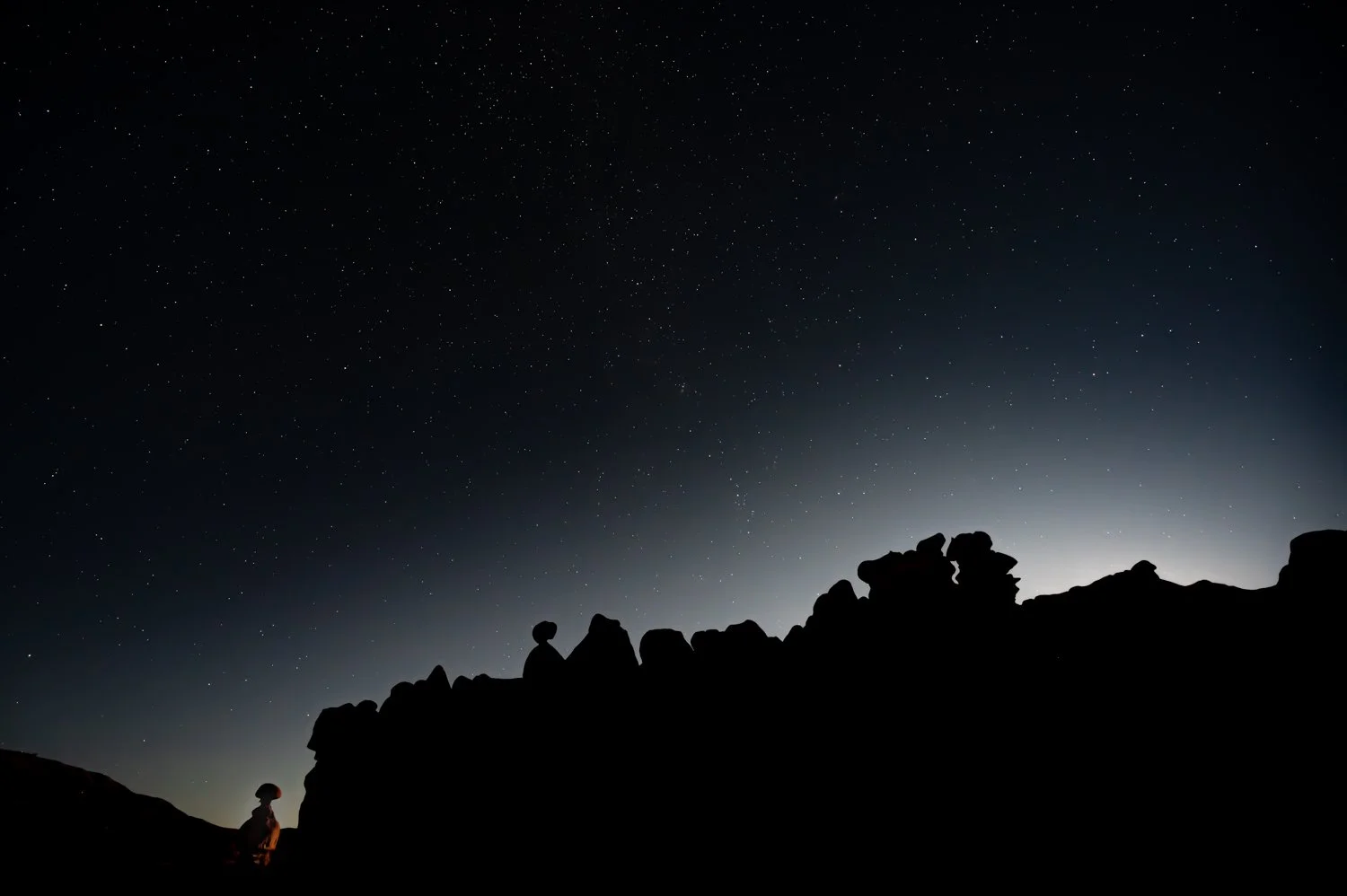 Silhouettes of hoodoos on rocks under a starry night sky with a bright glow near the horizon.