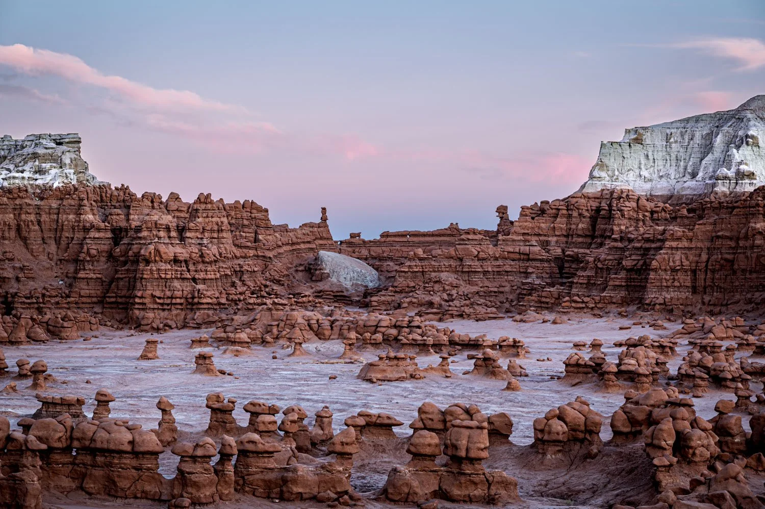 A desert landscape features various rock formations, including hoodoos and cliffs, under a pastel-colored sky with soft pink and blue hues at Goblin Valley State Park.