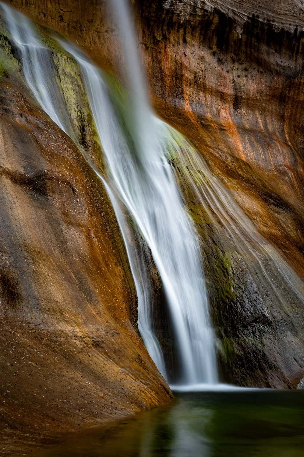 Water flowing down a narrow canyon with smooth, reddish-brown rock walls and a small pool at the bottom.