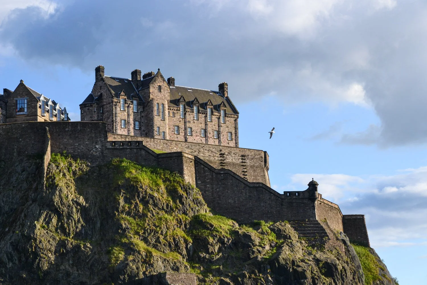 A large historic Edinburgh Castle with stone walls sits atop a rocky hill, with green grass and stairs leading up. The sky is partly cloudy and a bird is flying nearby.