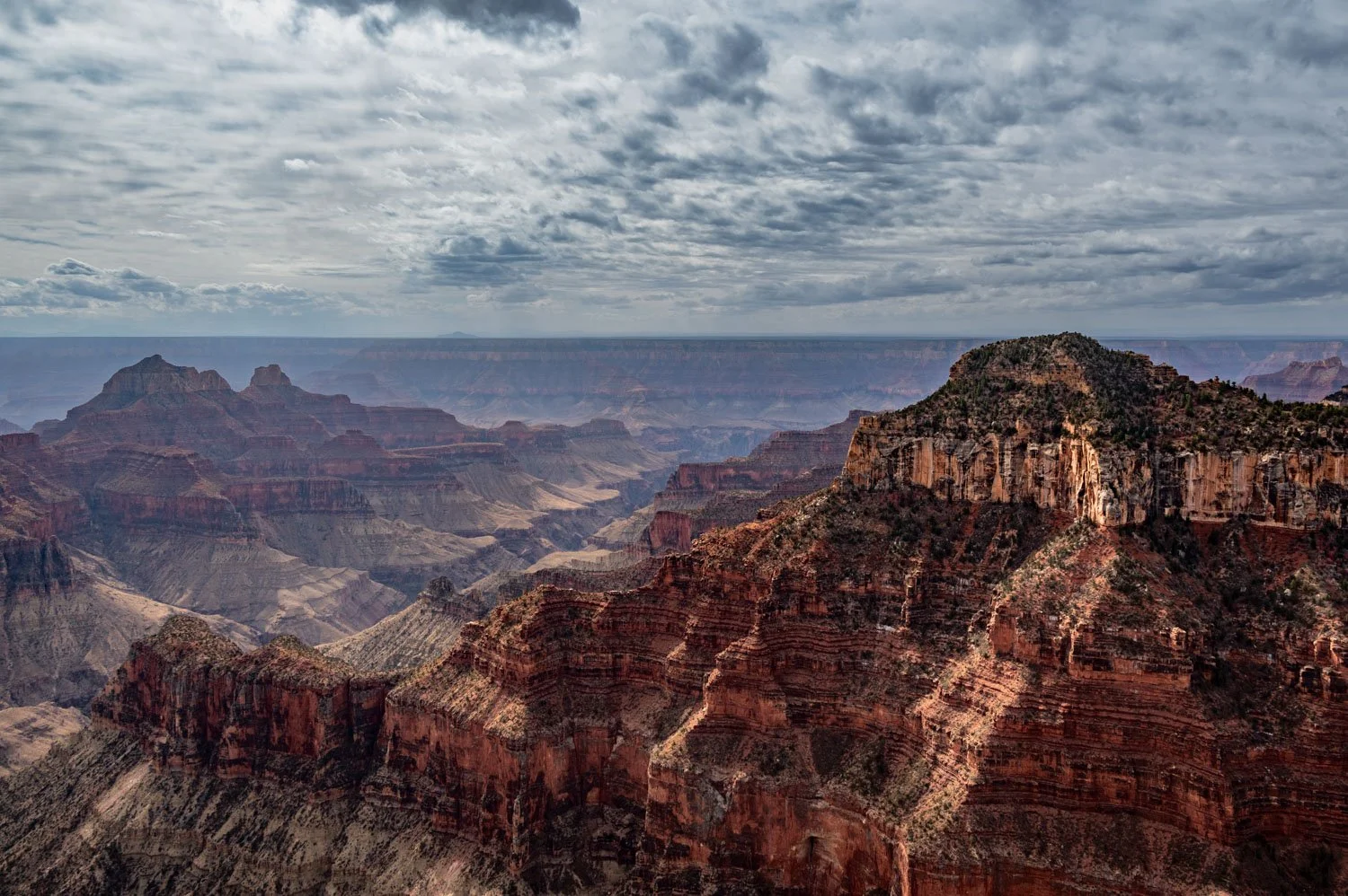 Panoramic view of the Grand Canyon with layered red rock formations and a cloudy sky.
