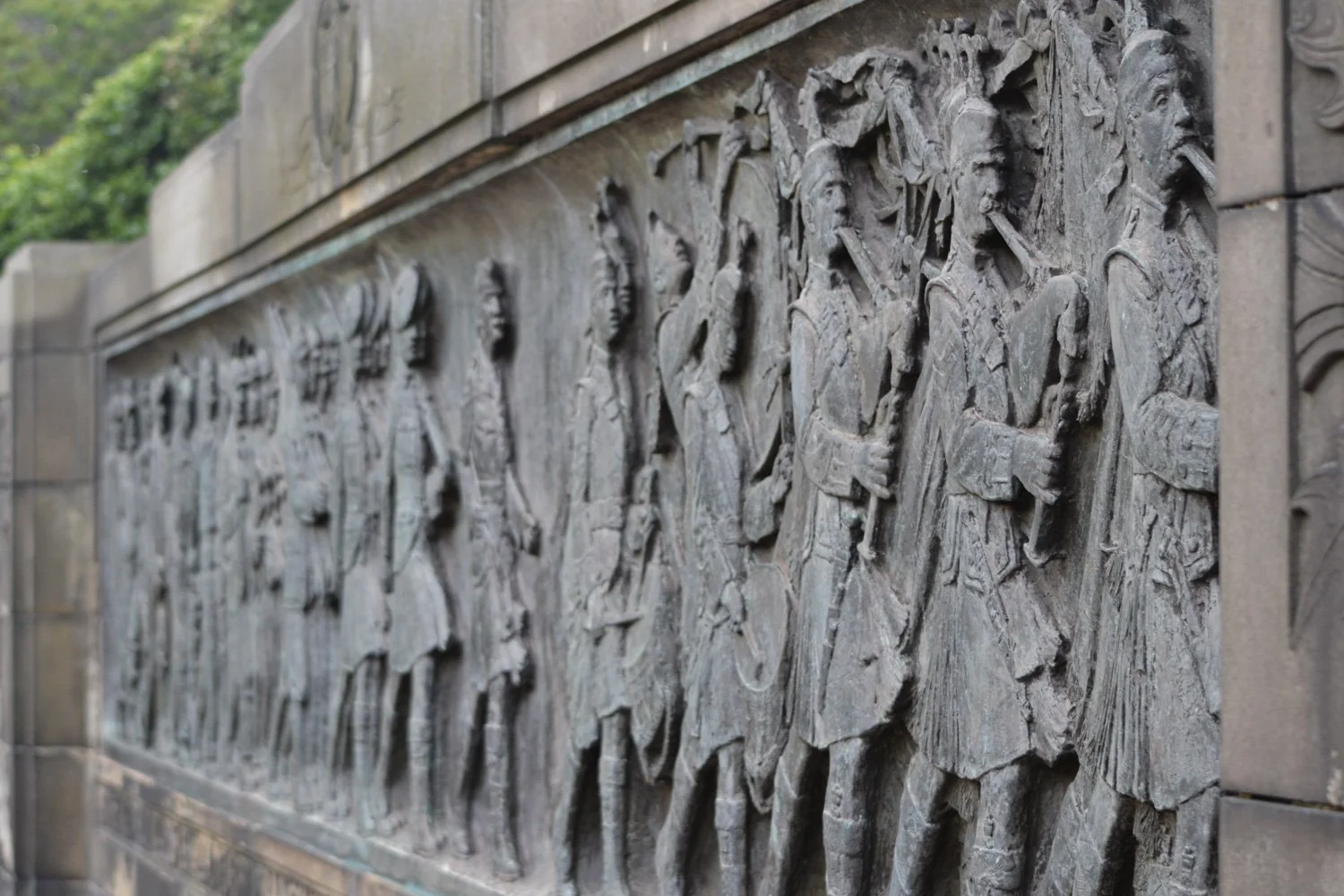 A long stone or metal relief sculpture depicting a group of historical or mythological figures, some carrying staffs or weapons, shown in profile.