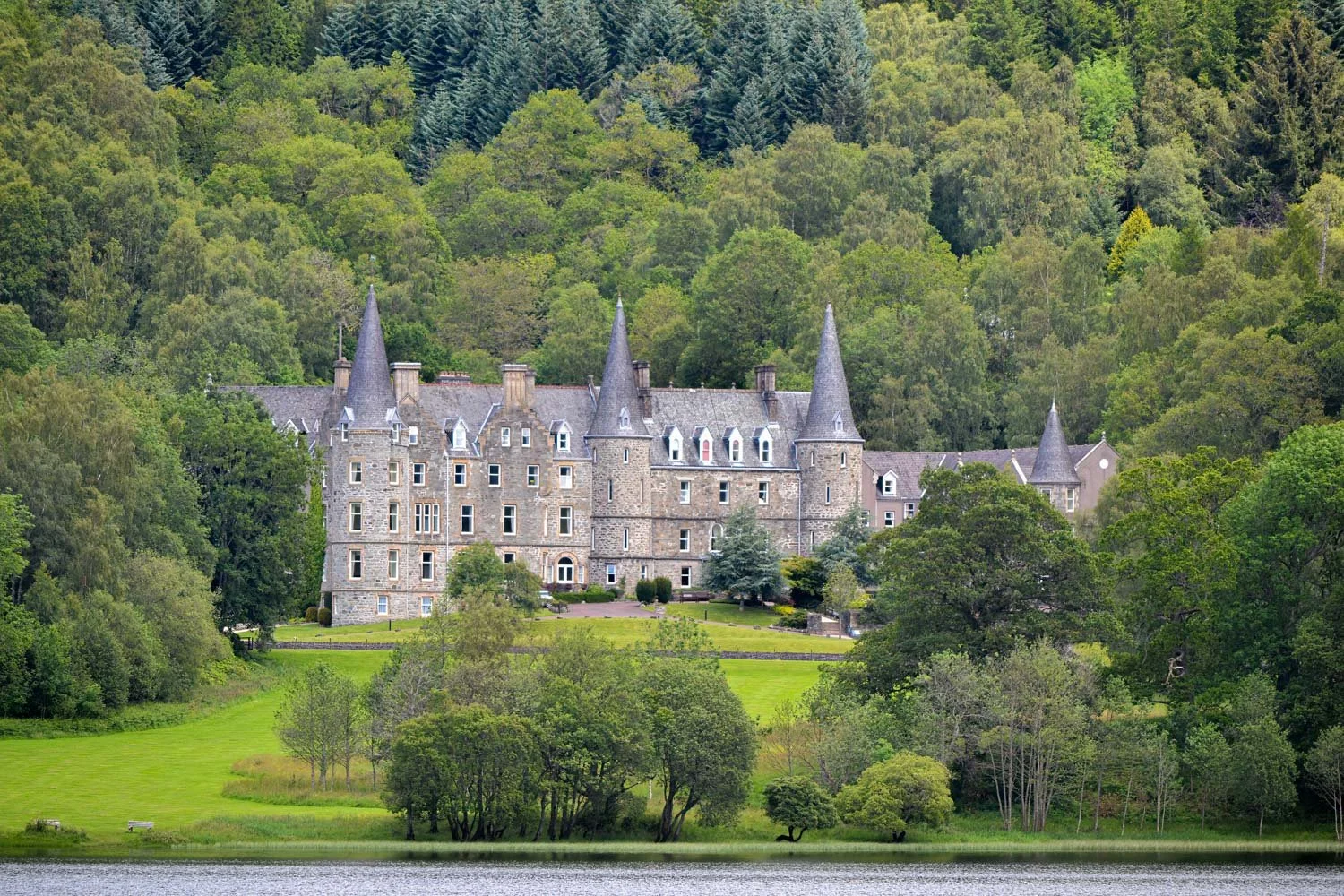 A large stone castle with pointed towers and turrets, surrounded by green trees and grass, near a body of water with a forested hillside in the background.