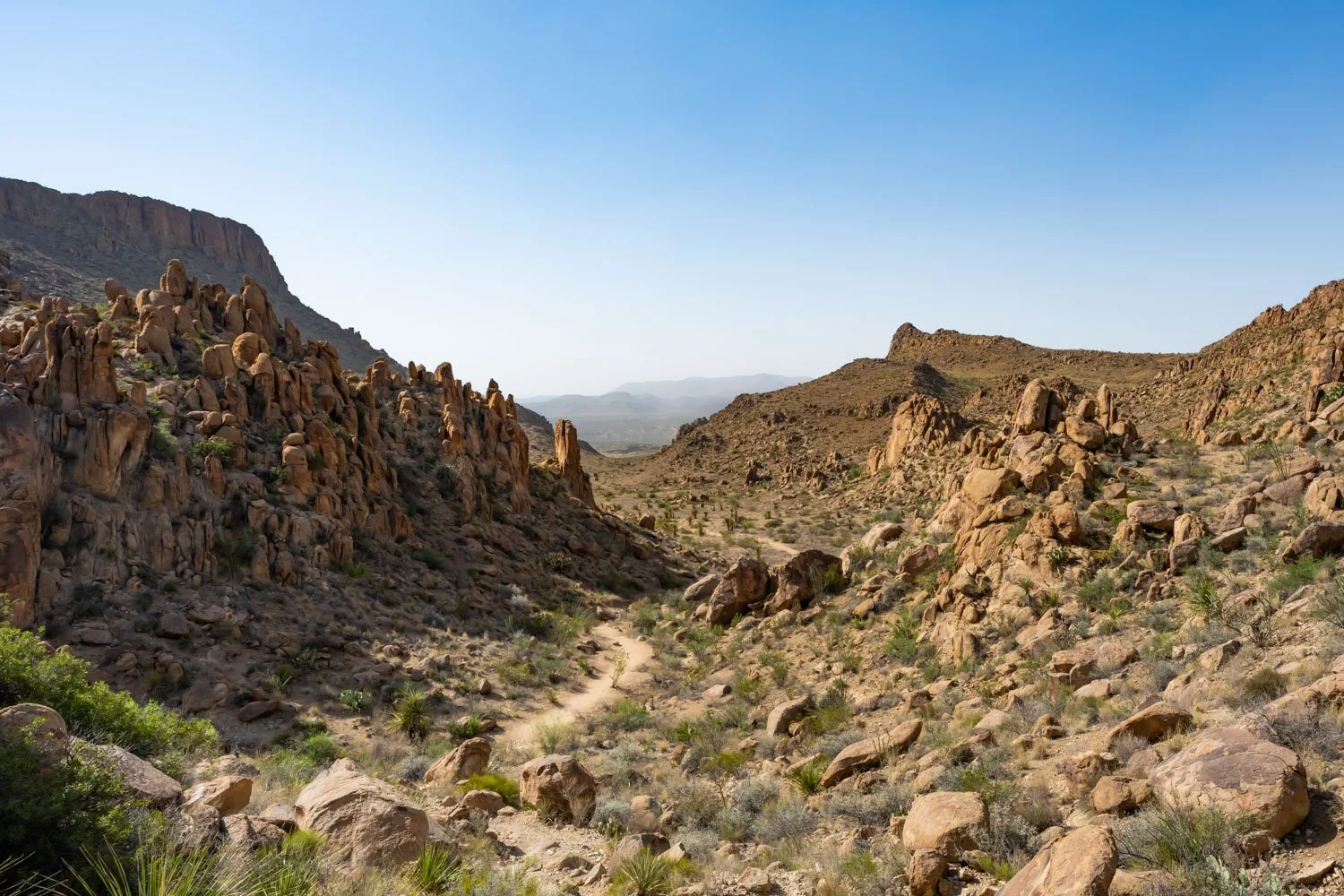A desert canyon landscape with rocky formations, sparse vegetation, and a dirt trail winding through the valley under a clear blue sky.