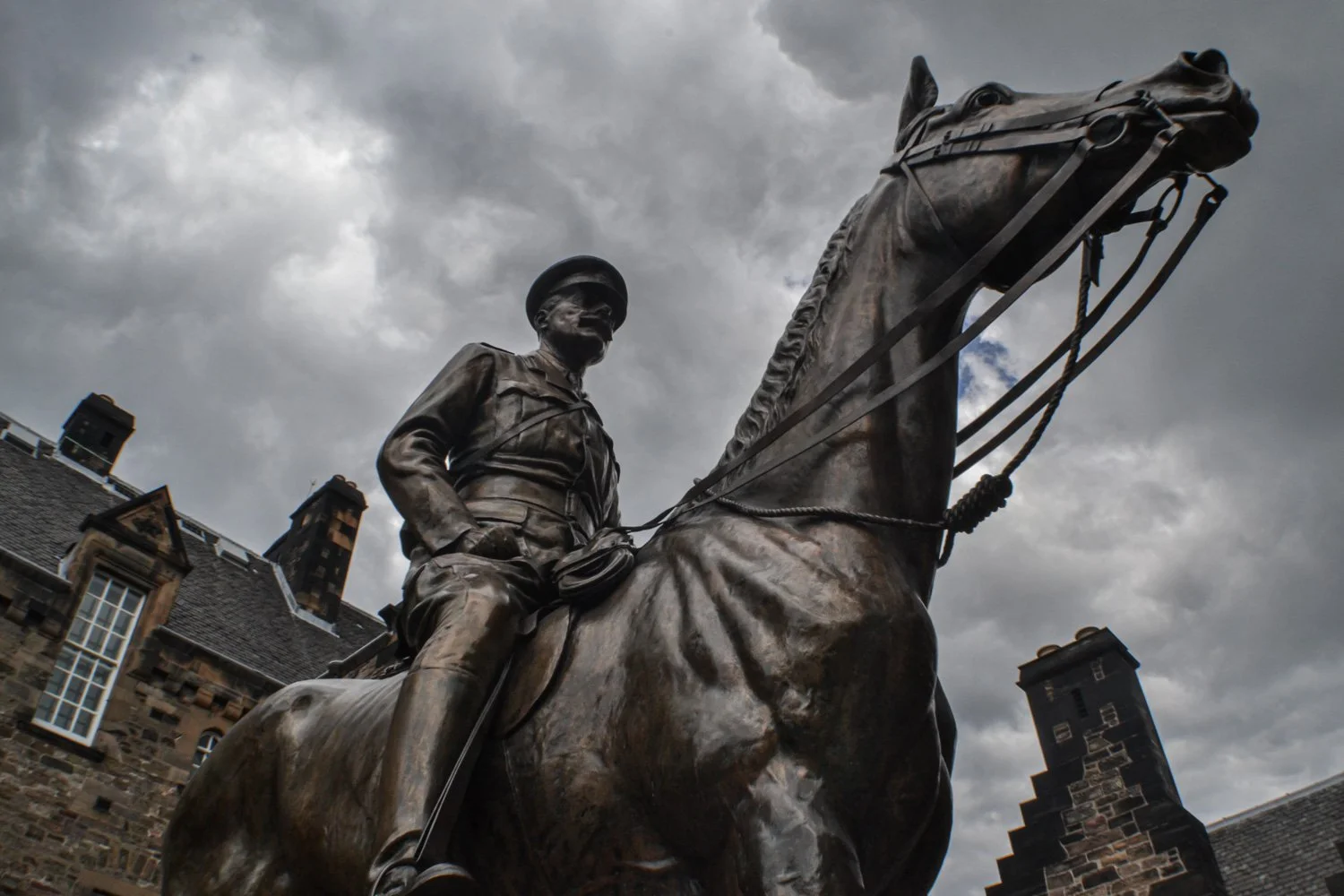 Bronze statue of a soldier on horseback under a cloudy sky, against historic stone buildings.