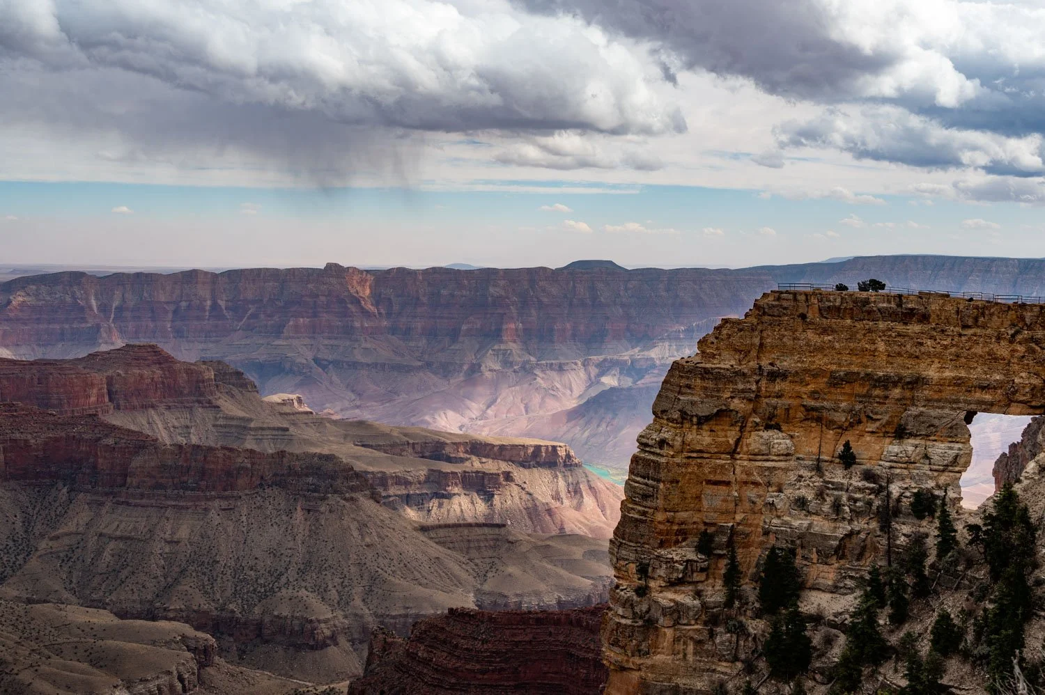 A panoramic view of the Grand Canyon with layered rock formations, a cloudy sky, and a small structure on top of a cliff.