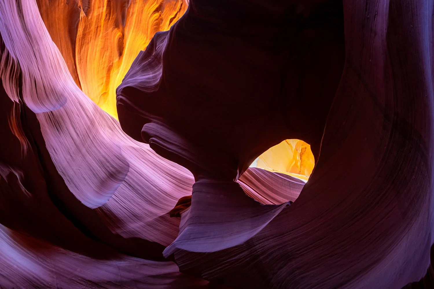 View of a colorful slot canyon with smooth, wavy rock formations in shades of purple, pink, orange, and yellow, with a small opening at the top letting in light.