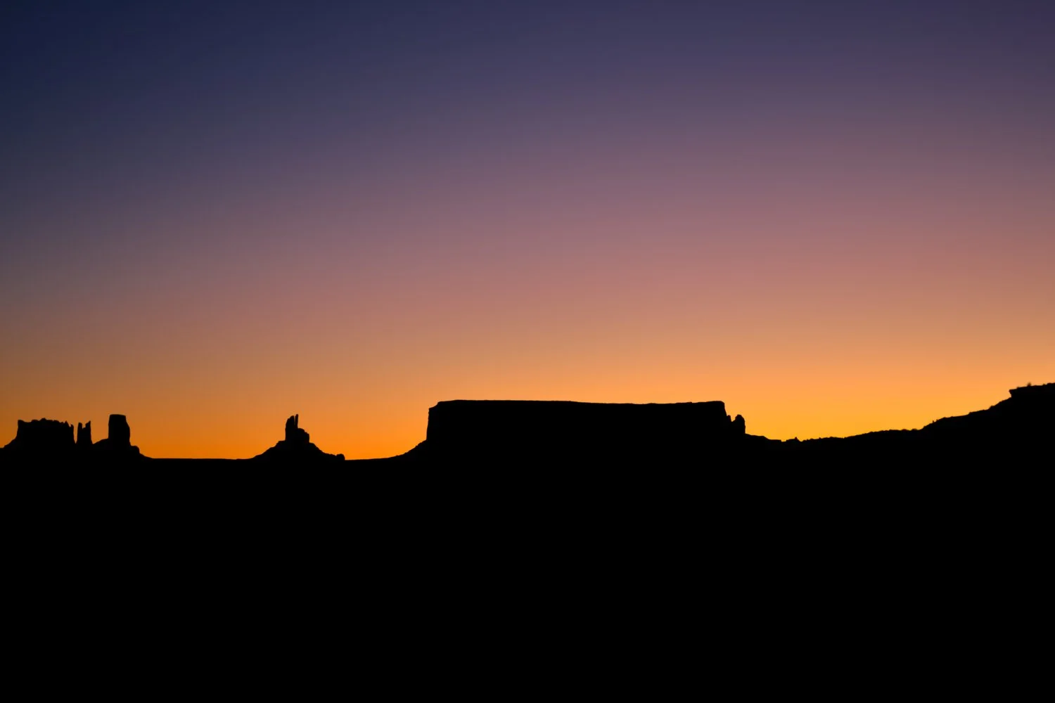 Silhouette of mesas and rock formations at Monument Valley against a colorful sunset sky with shades of purple, pink, and orange.