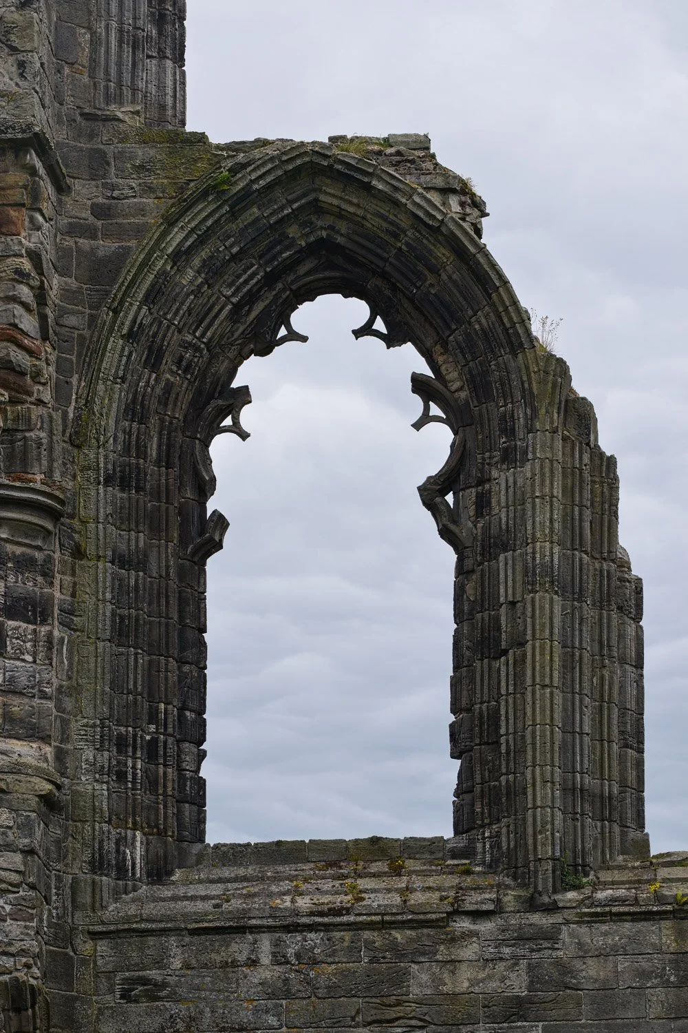 Ruined stone archway with gothic design, part of an old stone building, against a cloudy sky.