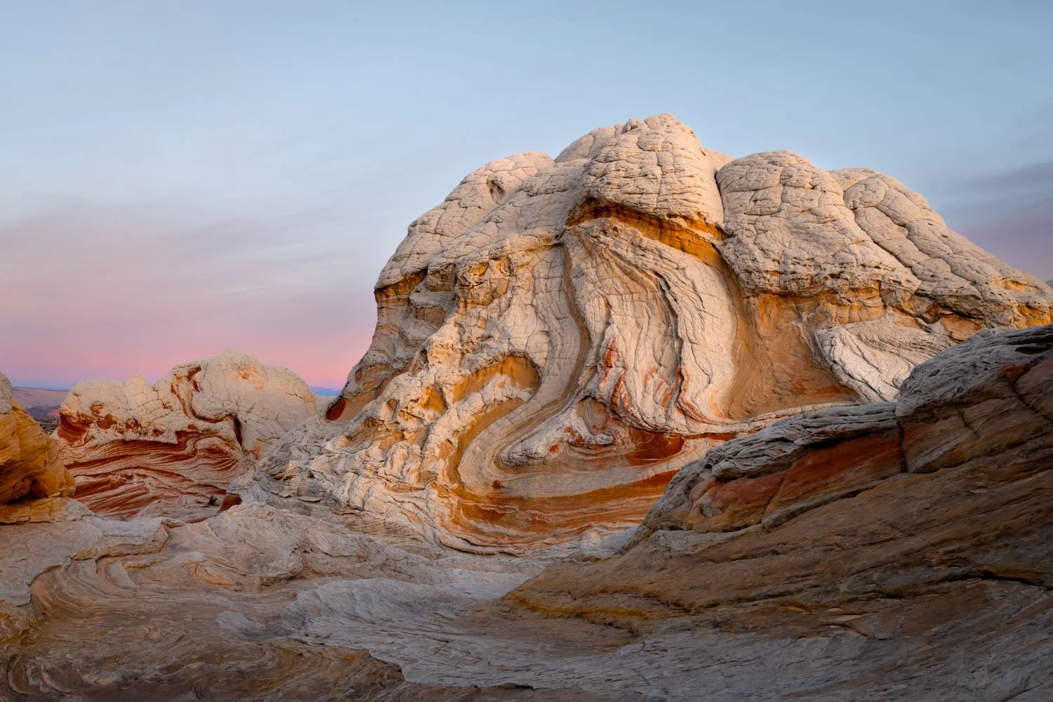 A large, uniquely shaped desert rock formation with swirling layers of white, cream, and reddish-brown colors, set against a pastel sky at sunset or sunrise.