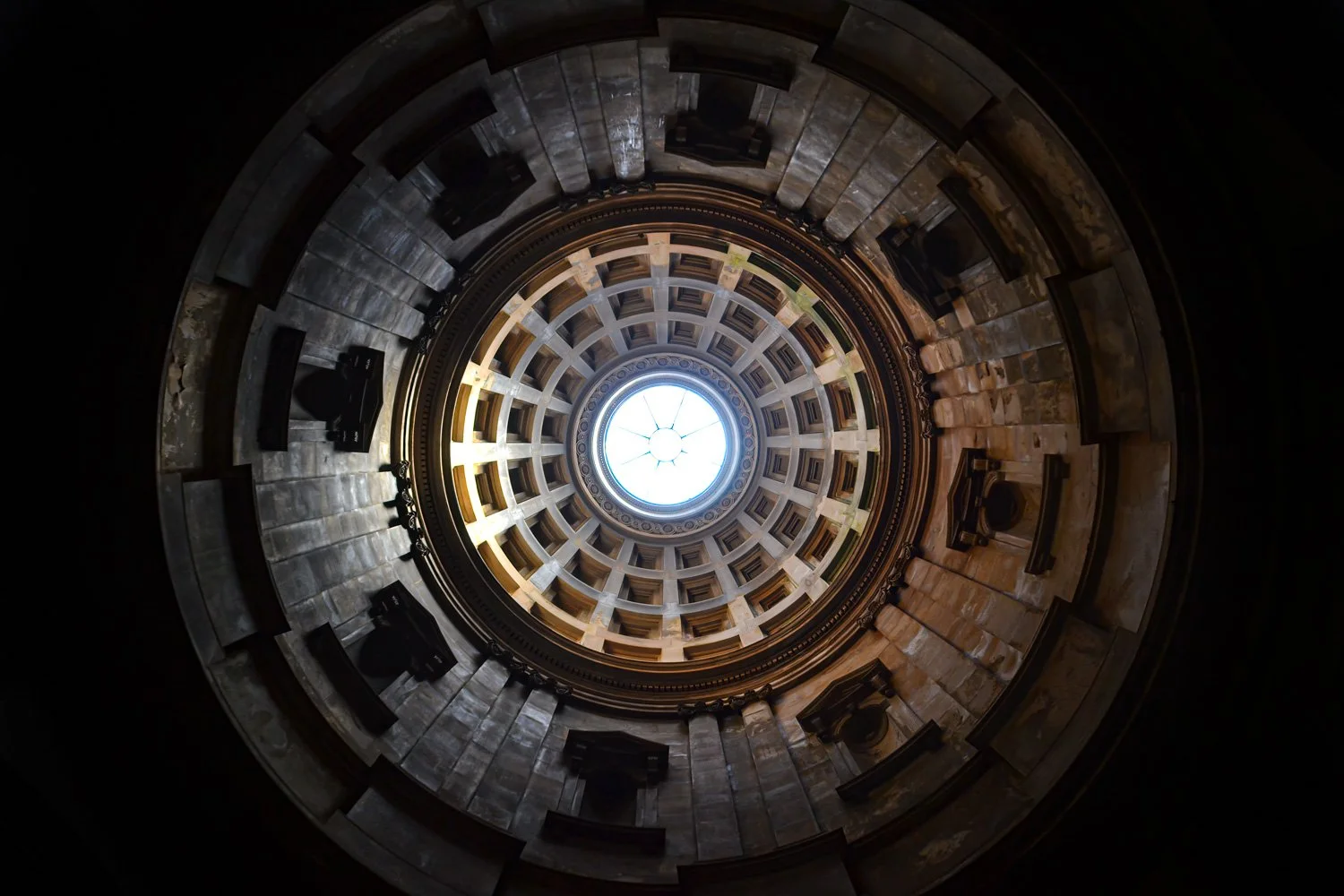 View looking up at the interior dome of a grand building with a circular glass skylight at the top, surrounded by multiple levels of architectural details.