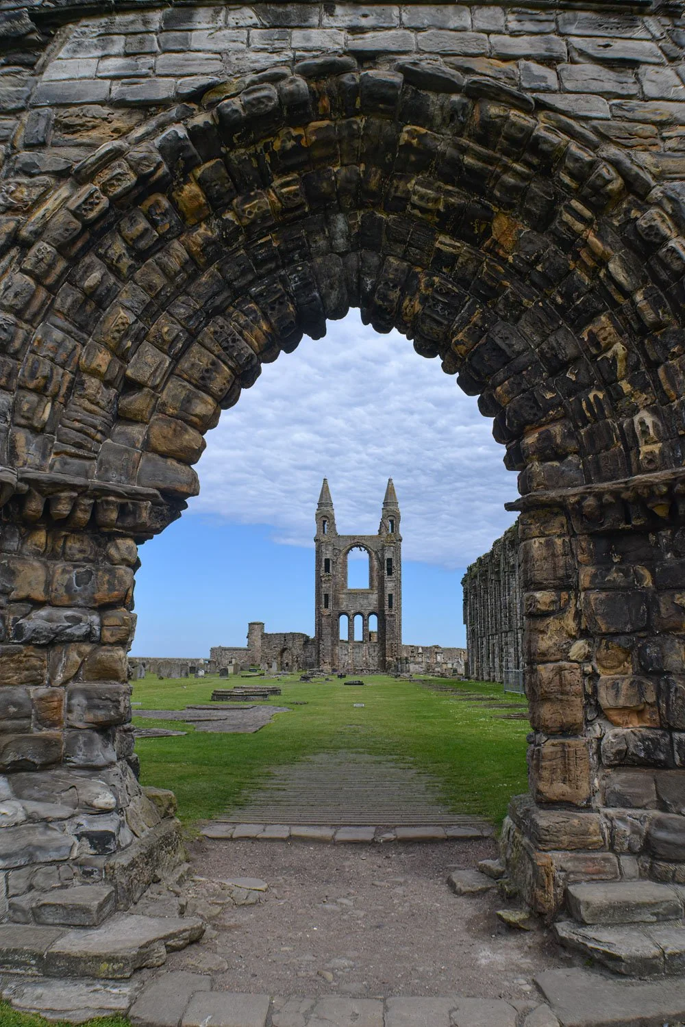 View through an ancient stone archway of the ruins of a historic church or cathedral, with a large tower featuring two spires in the distance under a cloudy sky.