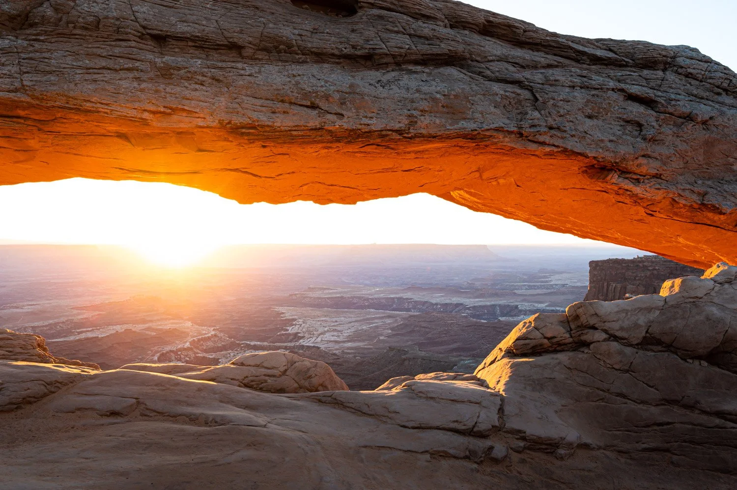 Sunrise view through a natural rock arch, overlooking a vast desert landscape with mesas and canyons.