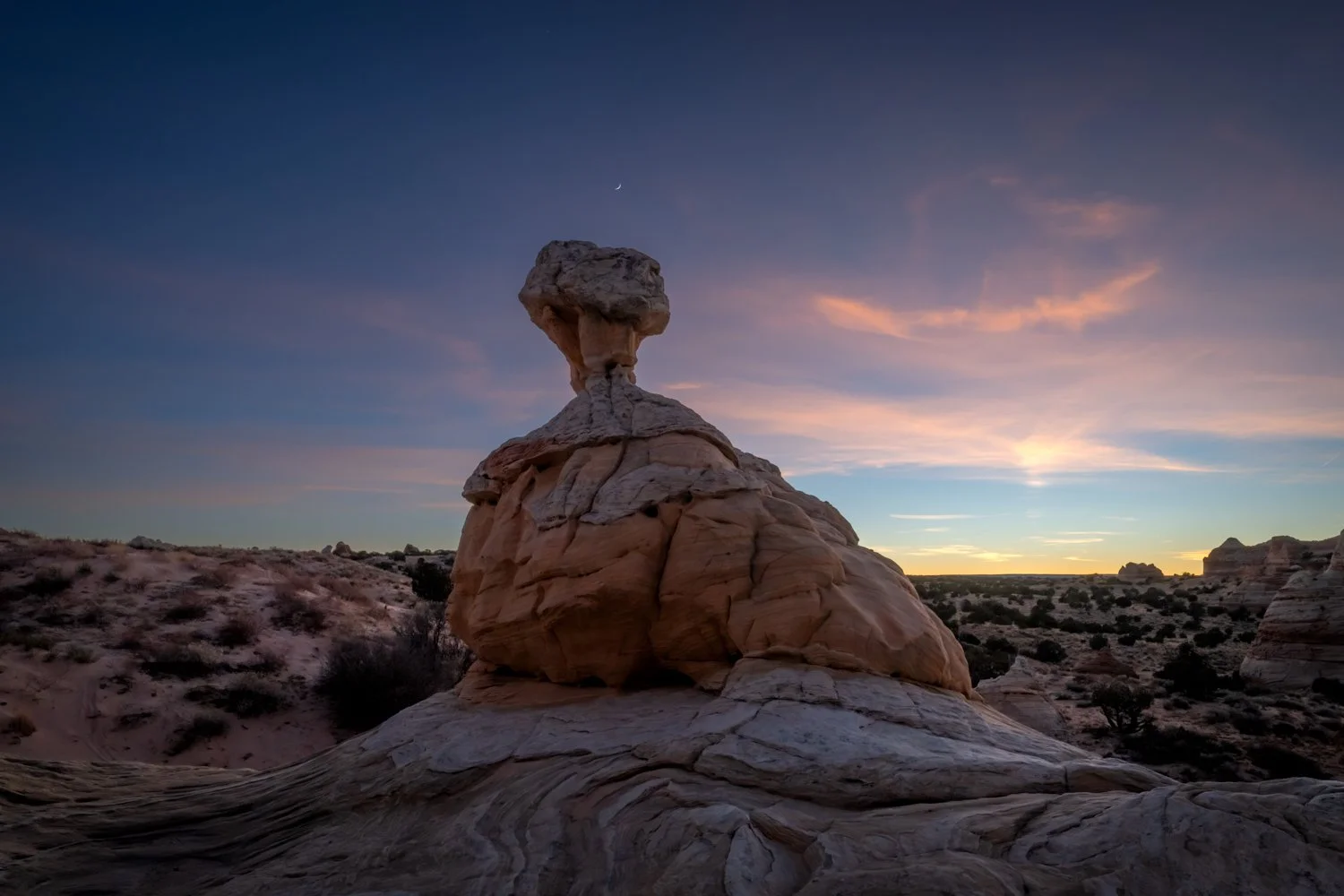 A desert landscape at sunset featuring a large rock formation with a top heavy shape, overlooking a vast desert with sparse vegetation and distant rock formations under a colorful sky with a crescent moon.