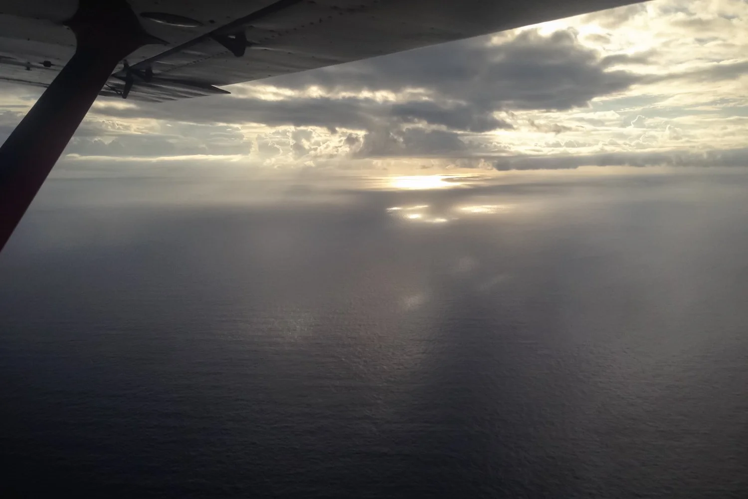 Aerial view from an airplane window showing the airplane wing, over the ocean with a partly cloudy sky and sunlight breaking through clouds.