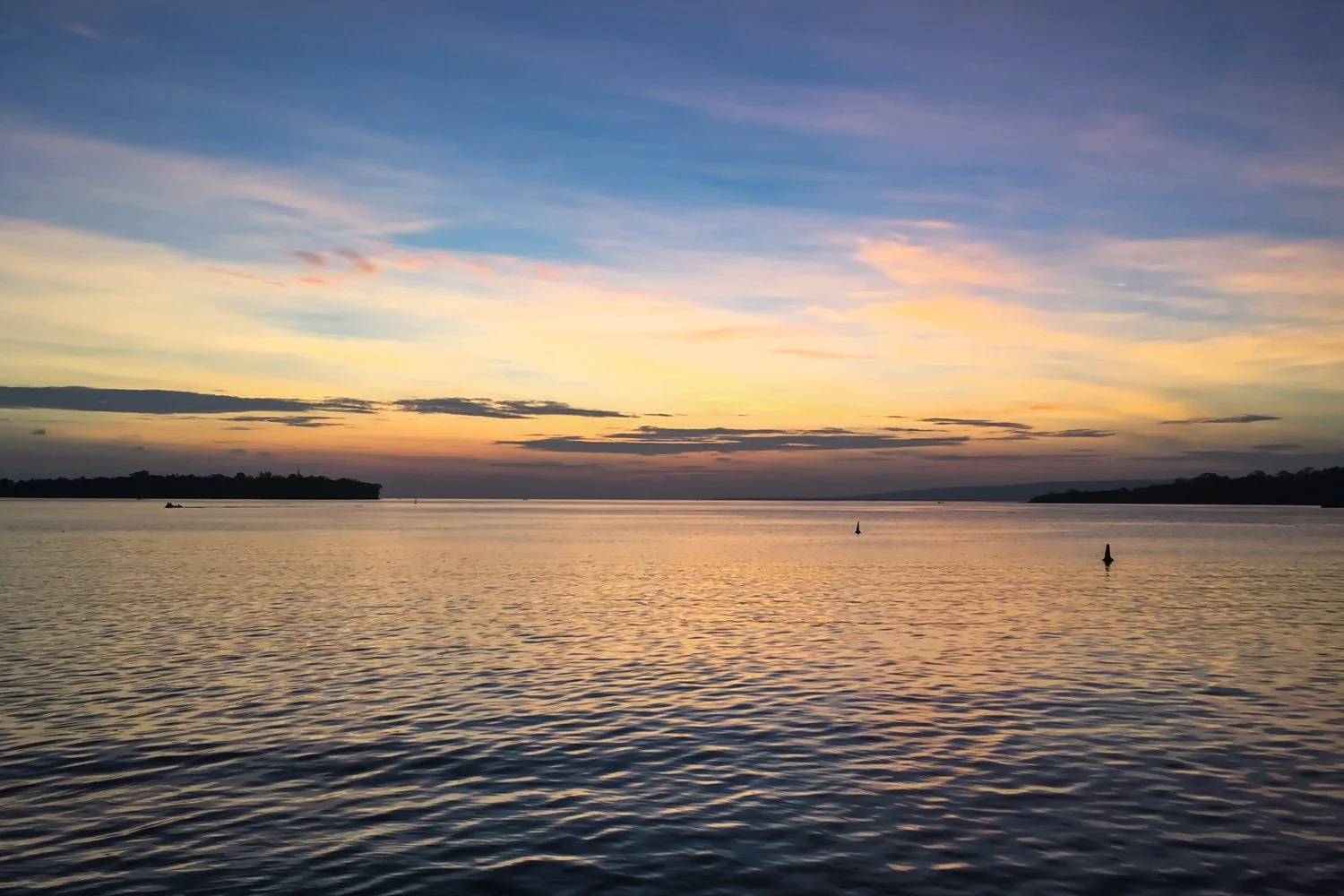 A serene body of water at sunset with calm ripples, distant treeline, and a colorful sky with shades of blue, pink, and orange.