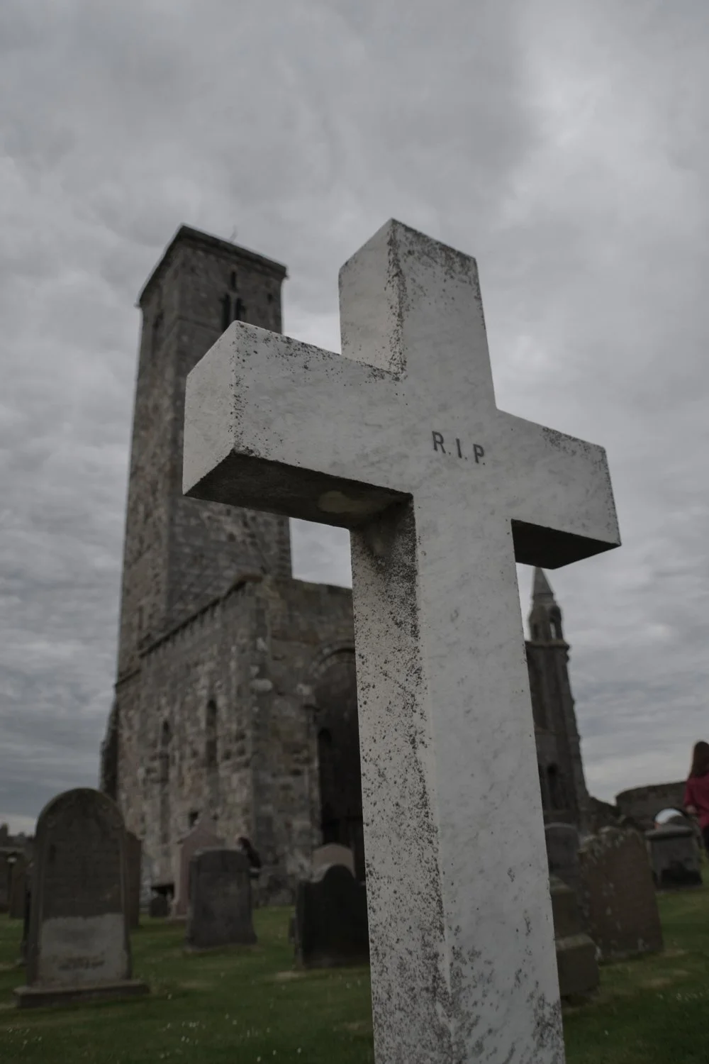 A white tombstone with 'R.I.P.' engraved on it, standing in a graveyard with a stone church and other graves in the background, under a cloudy sky.