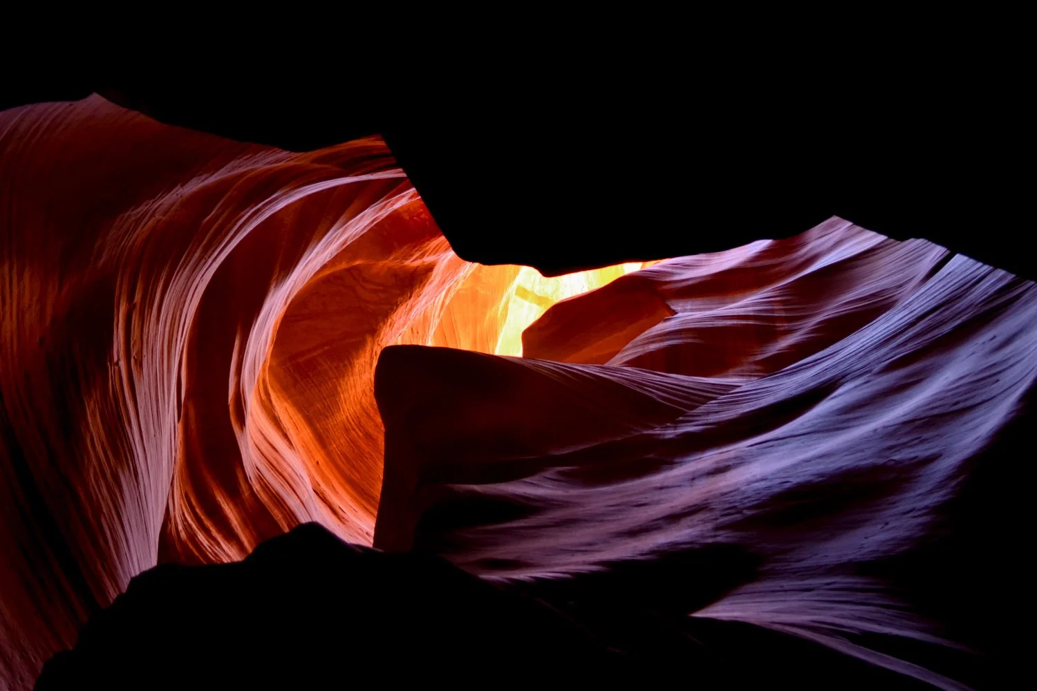 A view of a narrow slot canyon with smooth, flowing rock formations illuminated by sunlight, showcasing shades of orange, purple, and red.