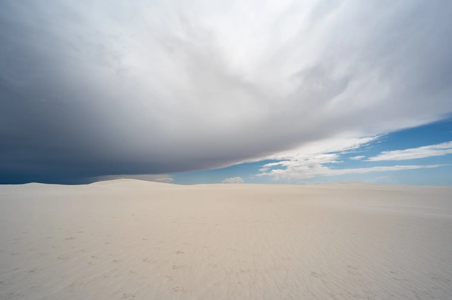 A wide view of a white sand desert under a large dark cloud with patches of blue sky and white clouds.