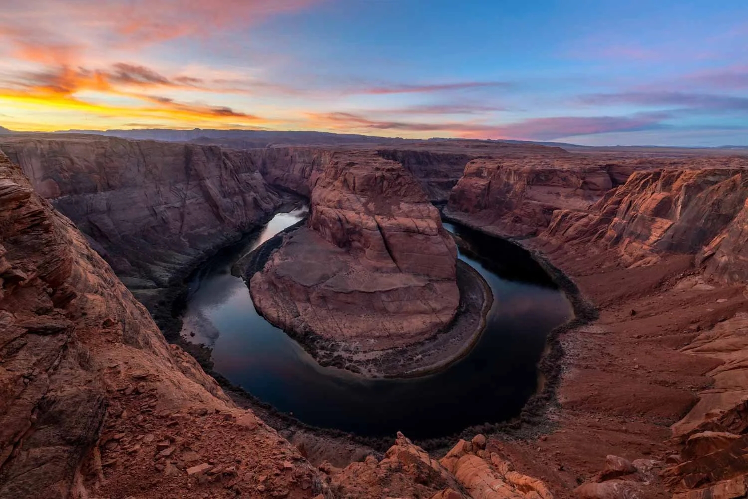 A panoramic view of Horseshoe Bend, a meander in the Colorado River, at sunset with a colorful sky and rugged canyon cliffs surrounding the river.