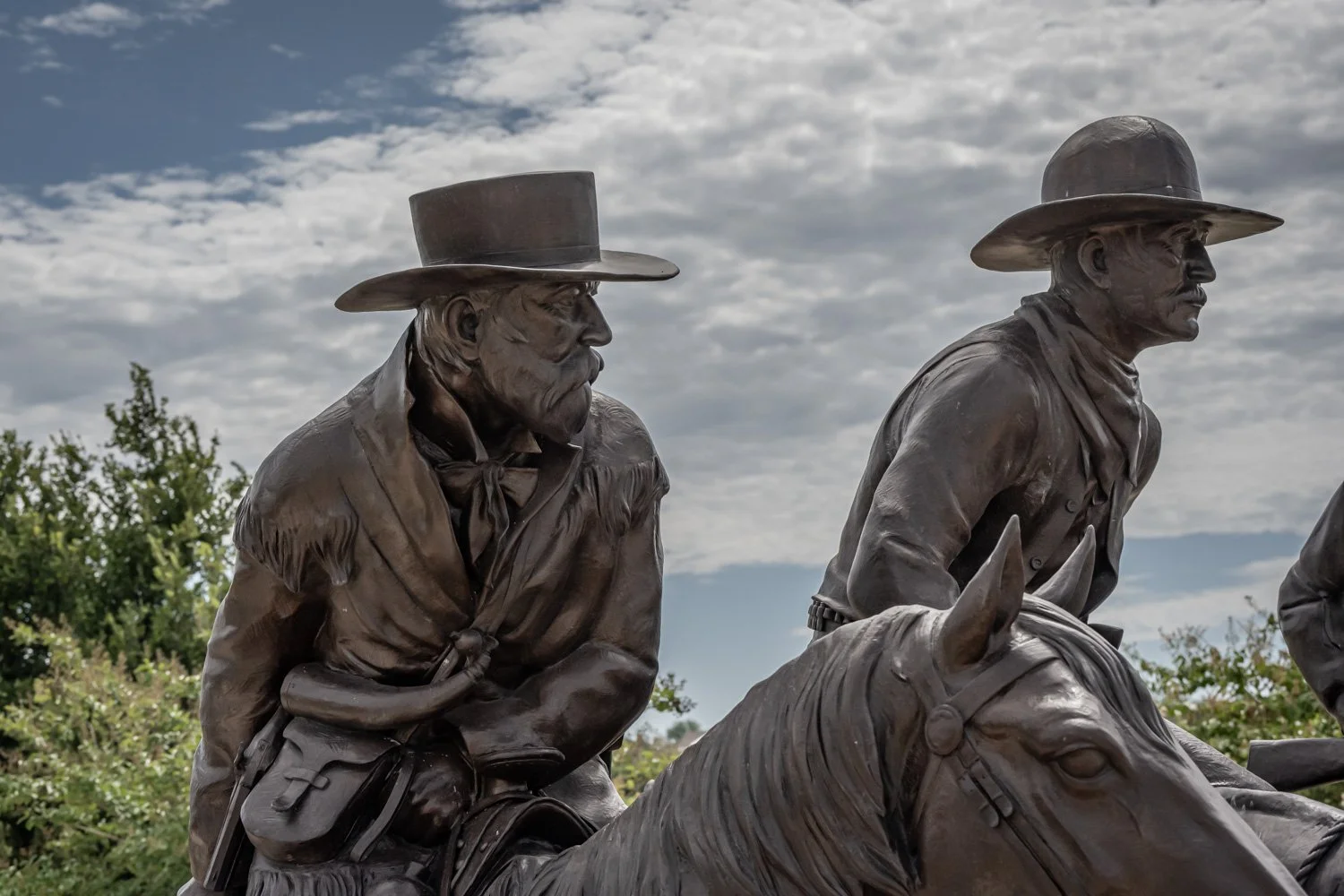 Bronze statues of two men in cowboy hats riding horses, set outdoors with cloudy sky and green bushes in the background.
