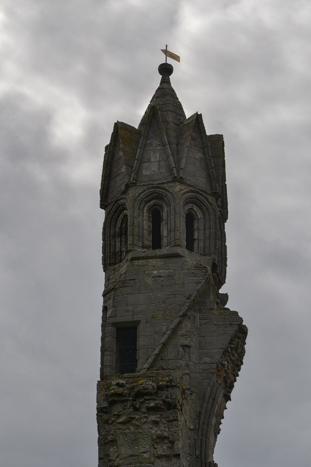 A tall, narrow stone tower with arched windows and a conical roof topped with a weather vane, against a cloudy sky.