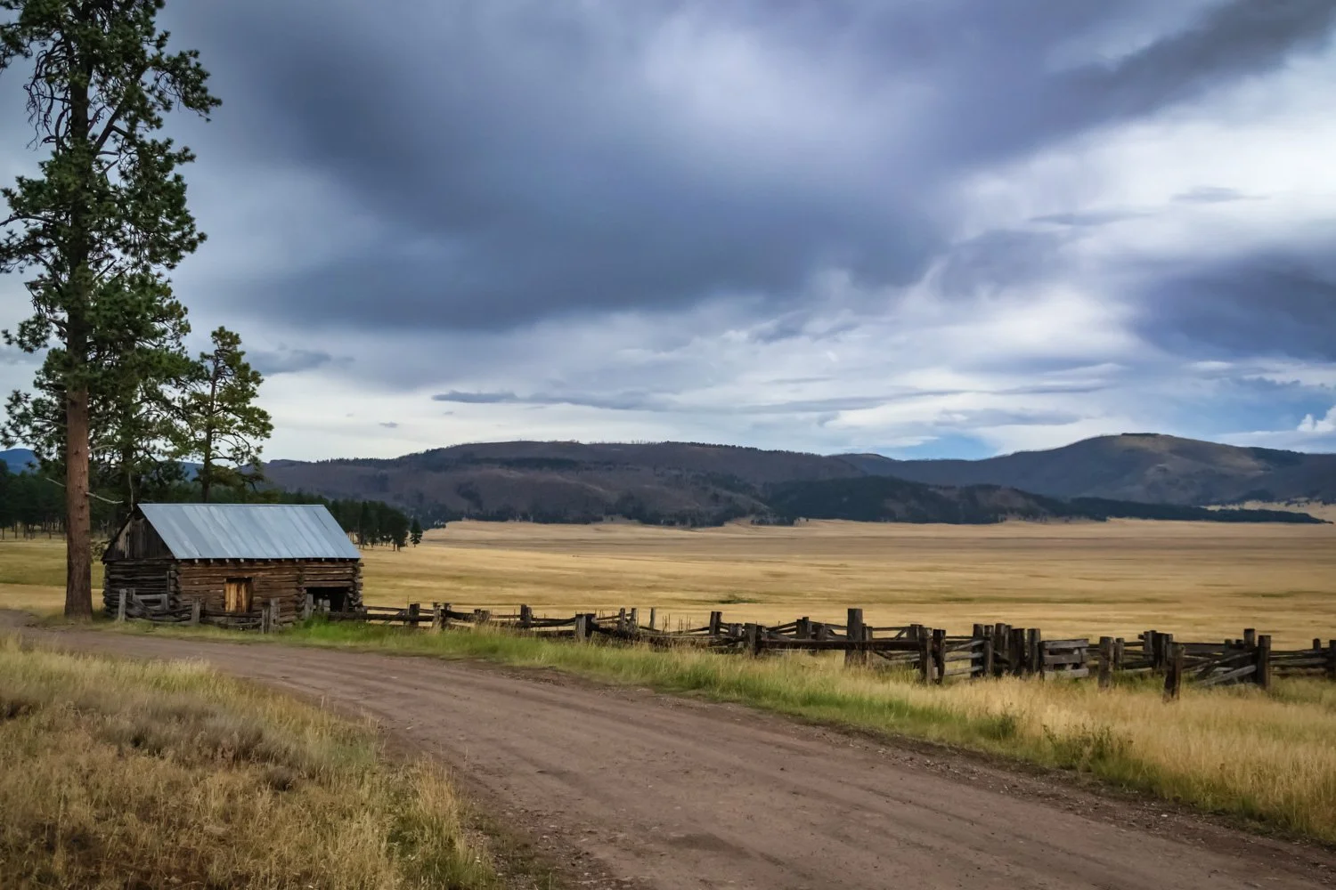 A rural landscape with a dirt road, an old wooden cabin with a metal roof, a wooden fence, grassy fields, pine trees, rolling hills, and dark cloudy sky.