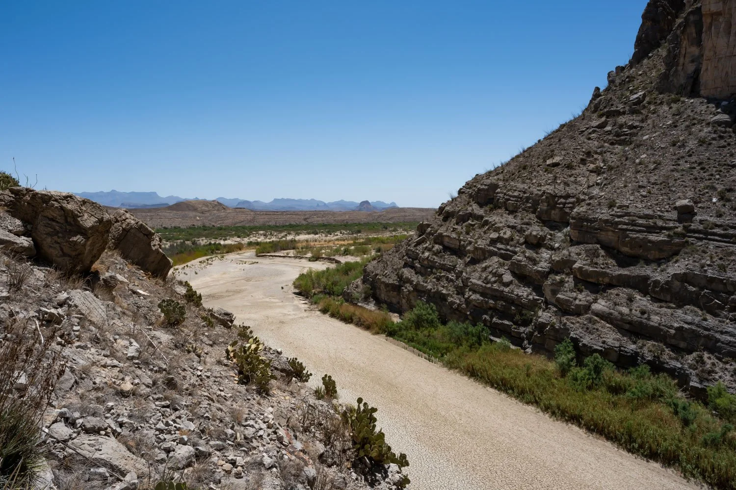 A dry riverbed of the Rio Grande winding through a rugged desert landscape with large rock formations and sparse vegetation under a clear blue sky.