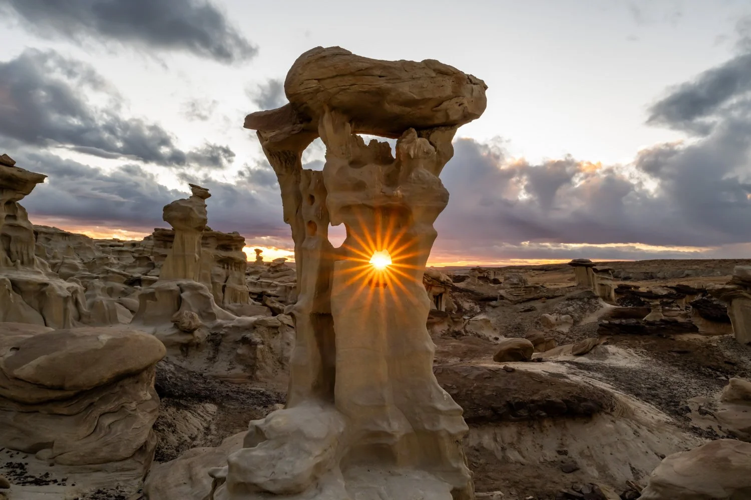 Sunset on a desert landscape with unique rock formations, with rays of the setting sun visible through a hole in one large rock formation called Alien Throne.