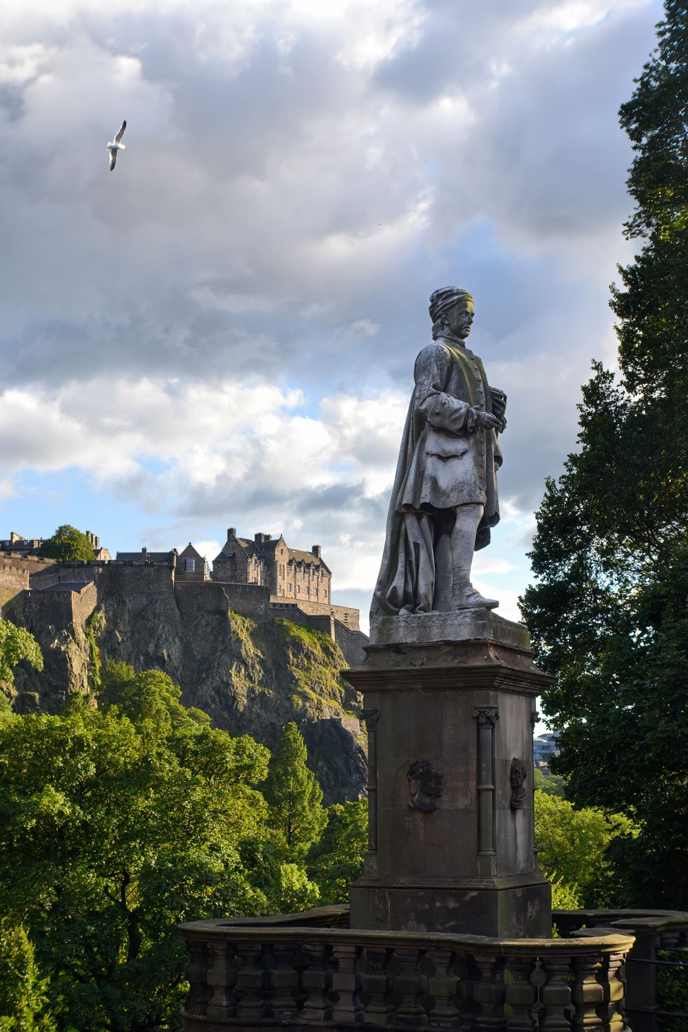 Statue of a historical figure on a pedestal in a park with Edinburgh Castle in the background and a seagull flying in the sky.
