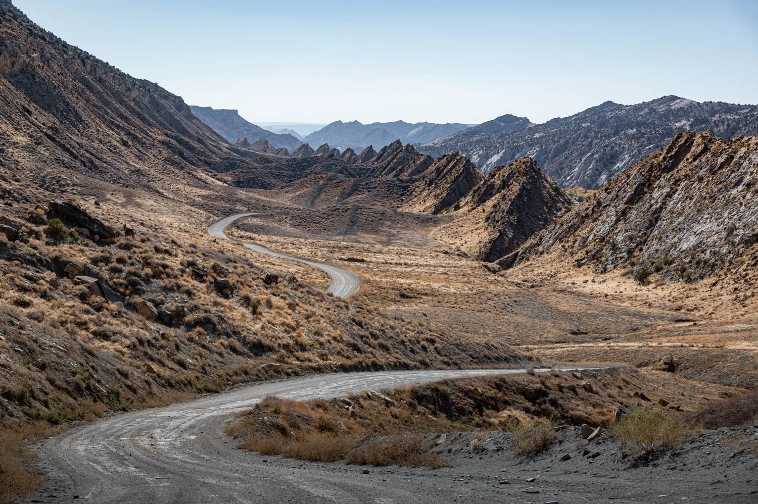 A winding dirt road runs through a dry, mountainous desert landscape with rugged, rocky hills on either side and minimal vegetation under a clear sky.