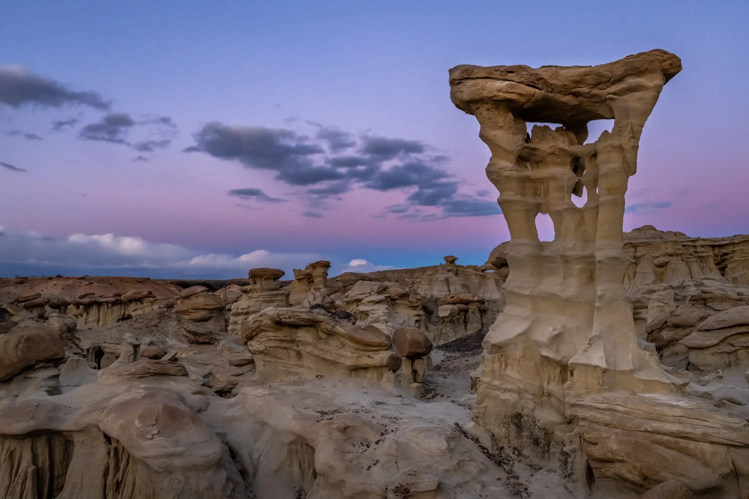 A desert landscape at dusk with unique rock formations, including a tall rock pillar called Alien Throne with holes and a flat top, under a sky with purple and blue hues and scattered clouds.