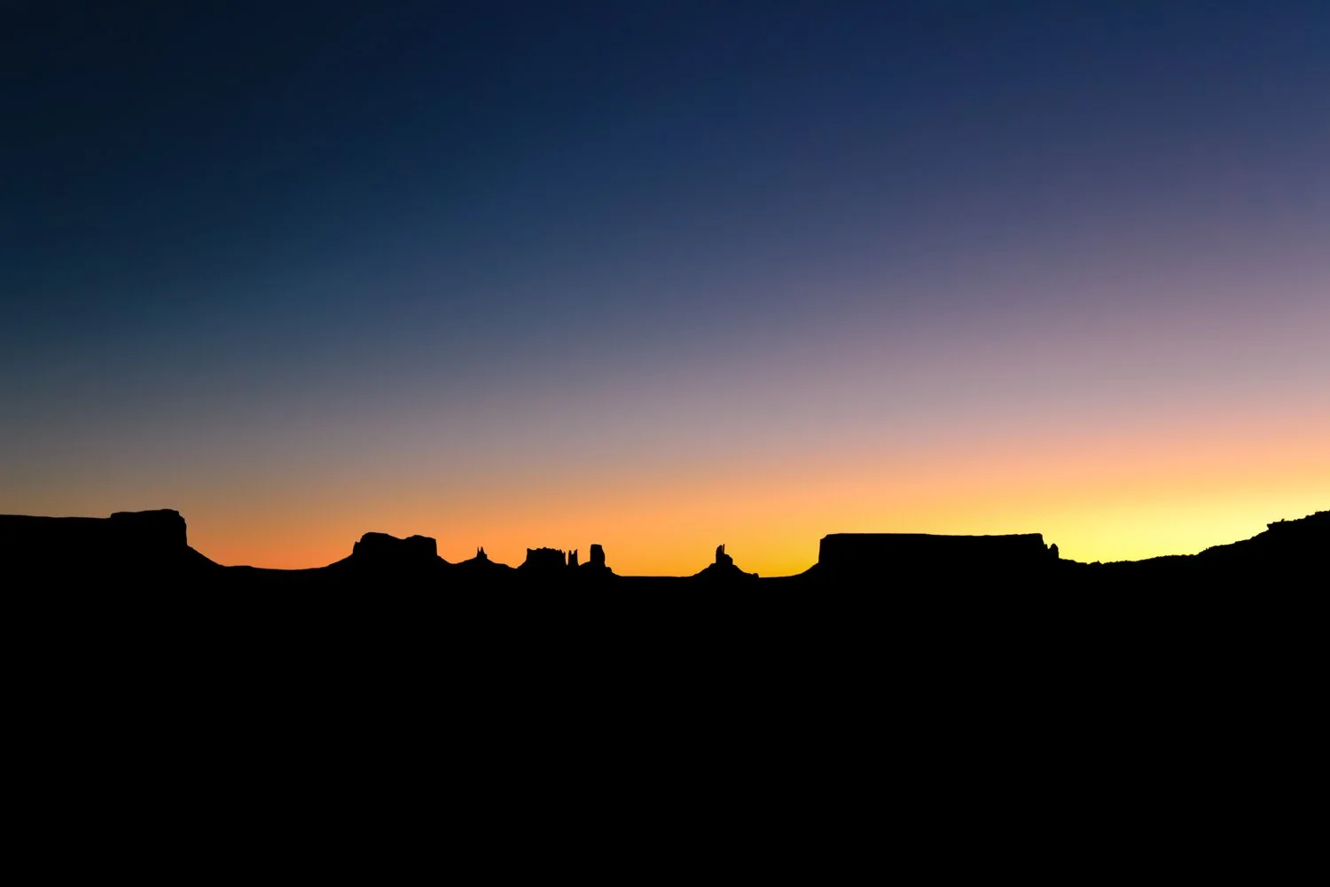Silhouette of a desert landscape at Monument Valley with rock formations against a colorful sunset sky transitioning from deep blue to orange.
