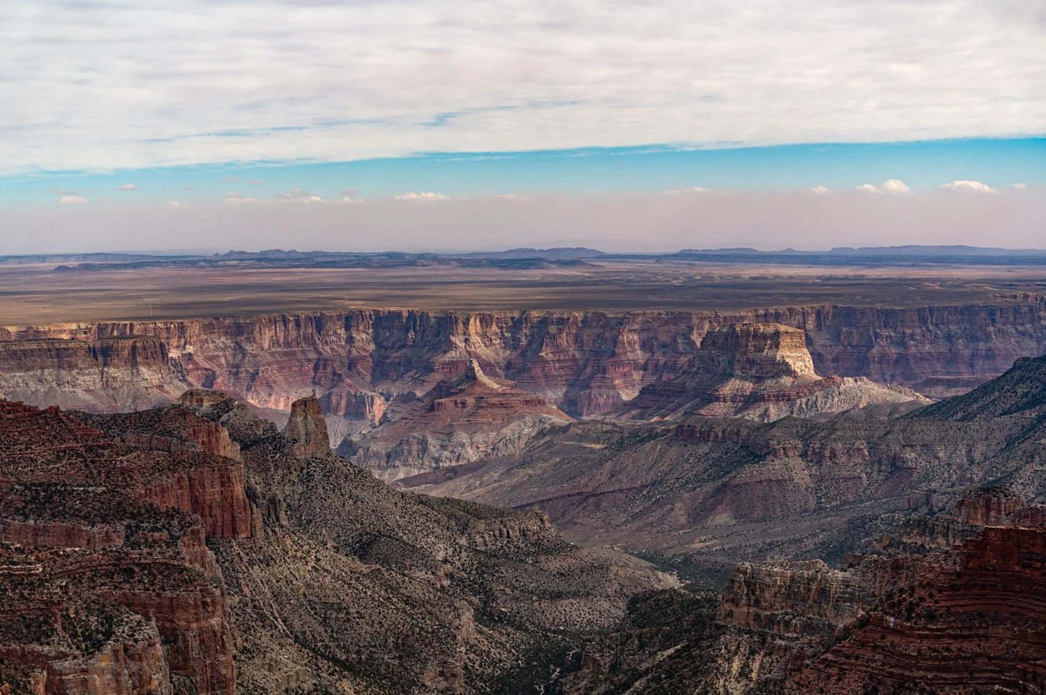A view of the Grand Canyon with layered rock formations, rugged cliffs, and a distant flat landscape under a partly cloudy sky.