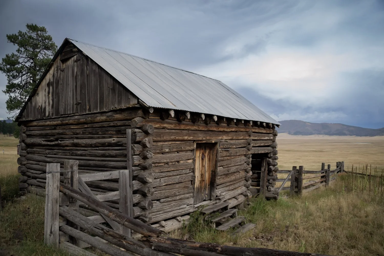 A rustic log cabin with a metal roof situated in a rural landscape, surrounded by grass, a wooden fence, and distant hills.