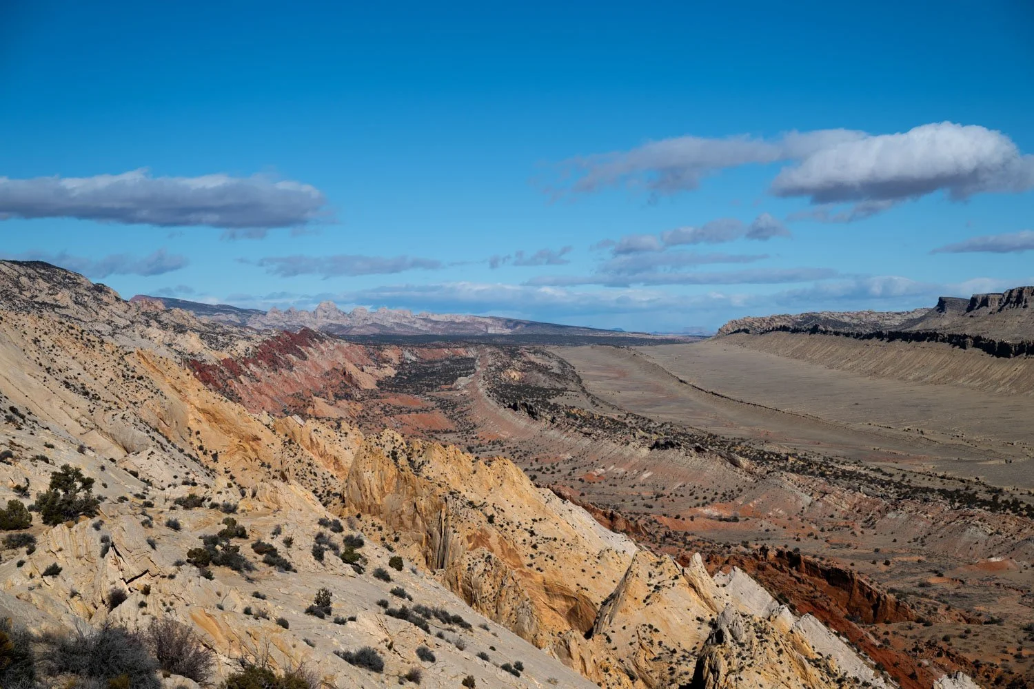 A scenic view of a vast desert canyon landscape of the Water Pocket Fold in Capitol Reef National Park with colorful rocky formations, sparse vegetation, and a partly cloudy sky.