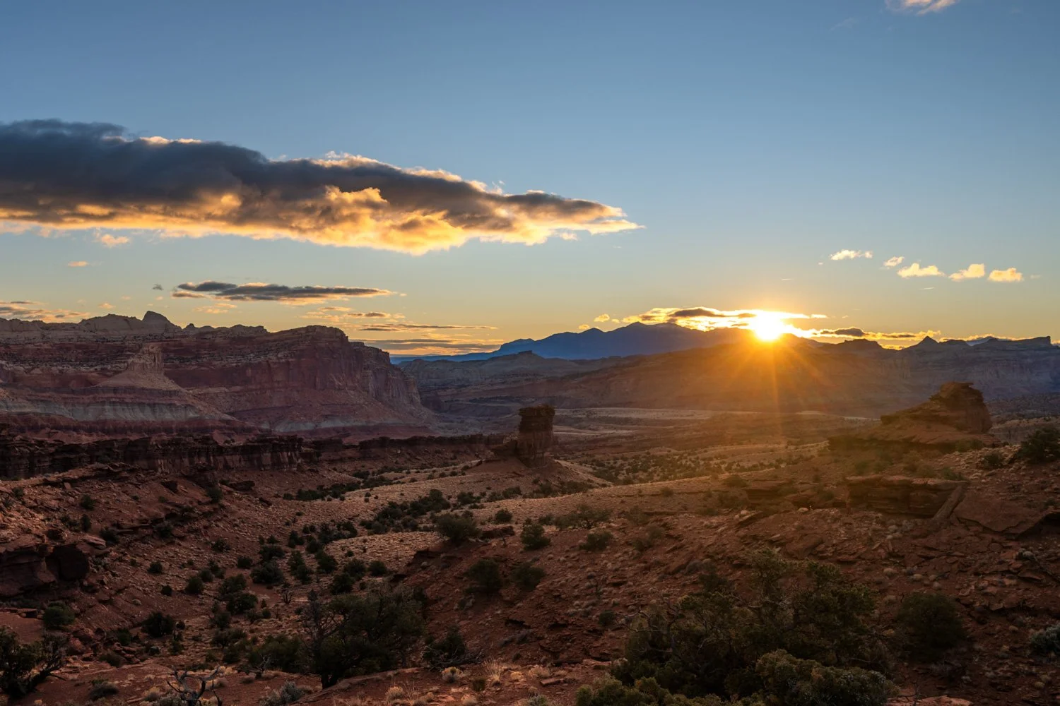 Sunrise over a desert canyon with distant mountains, rock formations, and sparse vegetation.