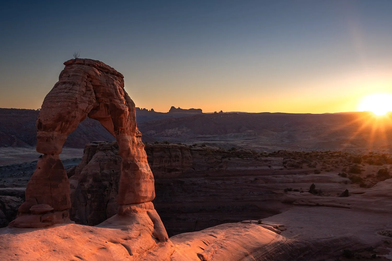 Arches National Park sunrise showing the Delicate Arch, a natural sandstone formation, with the sun setting over a desert landscape of rock formations and canyons.