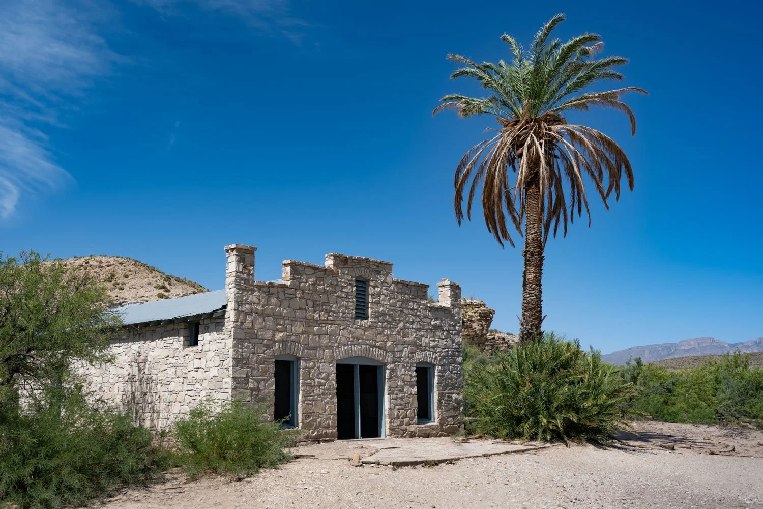 An old stone building with a metal roof next to a tall palm tree in a desert landscape with mountain background.