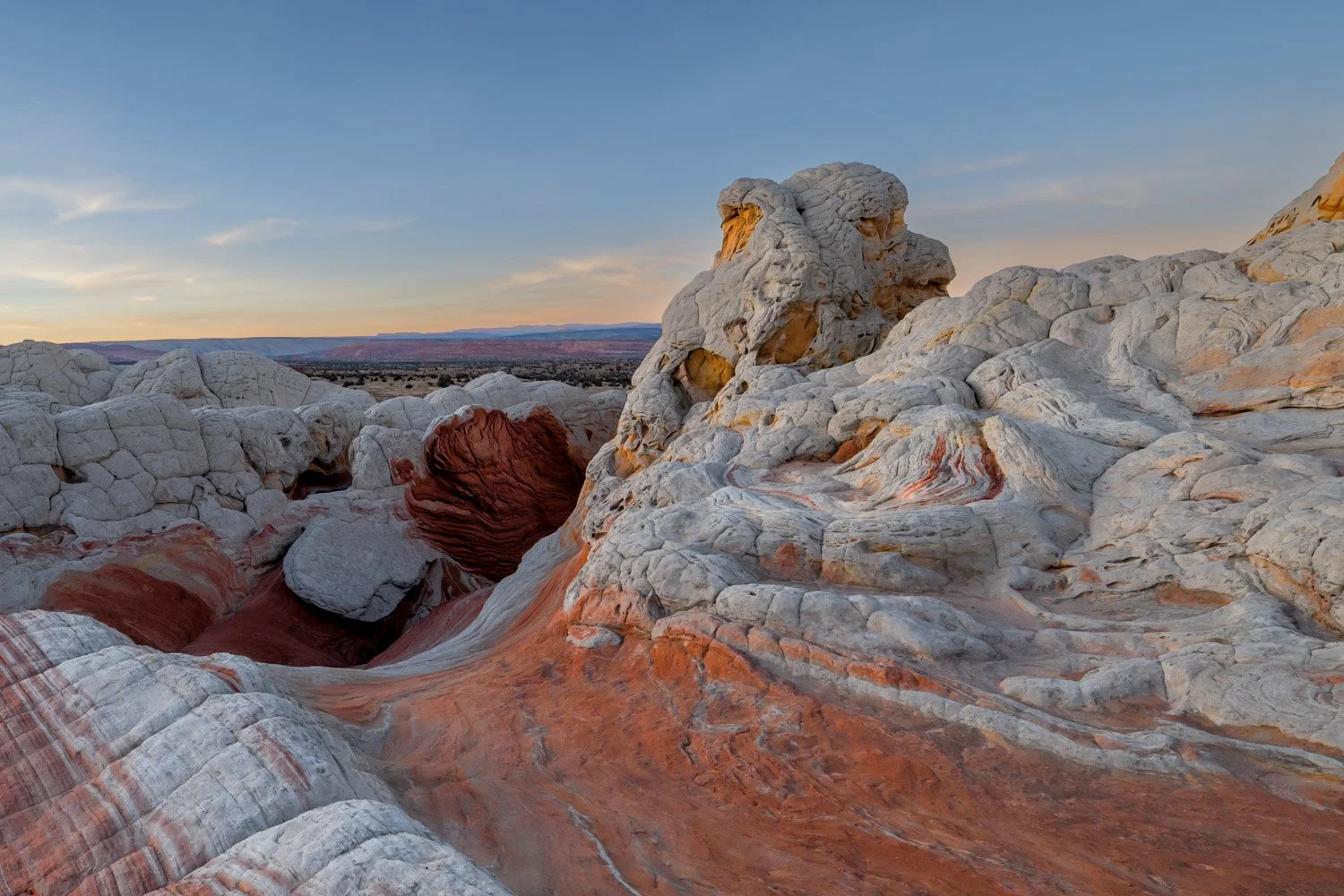 Desert landscape with multicolored rock formations, including white and reddish layers, under a clear sky at sunset.