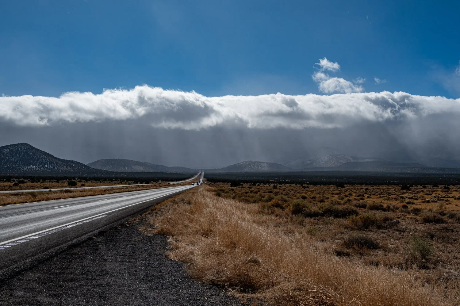 A long, straight highway running through an arid landscape with low shrubs, leading towards distant mountains under a cloudy sky with patches of blue.